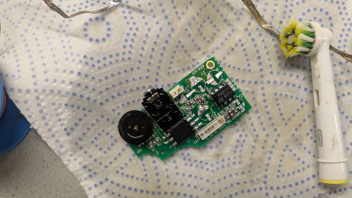 Game Gear audio/power board after ultrasonic cleaning, laid on dot matrix mat with soldering iron and yellow-flowered plant visible.