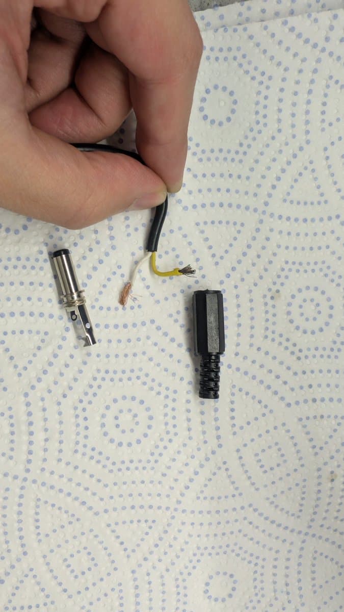 Disassembled Game Gear external PSU connector showing separated barrel jack, cable with exposed yellow and black wires, and connector housing on dotted fabric background.