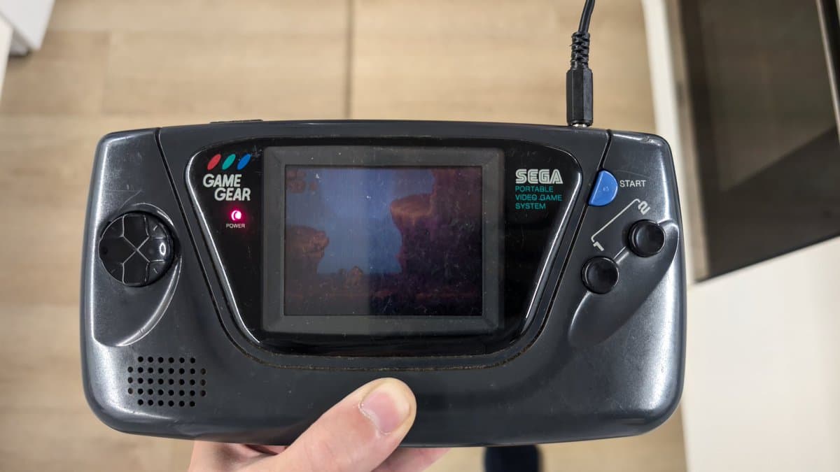 Sega Game Gear held in hand displaying repaired screen with underwater scene, power LED lit, external PSU restored.