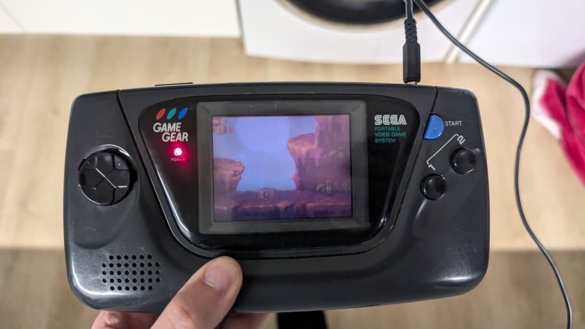 Sega Game Gear displaying colorful desert landscape game scene on screen, held in hand showing successful repair completion.