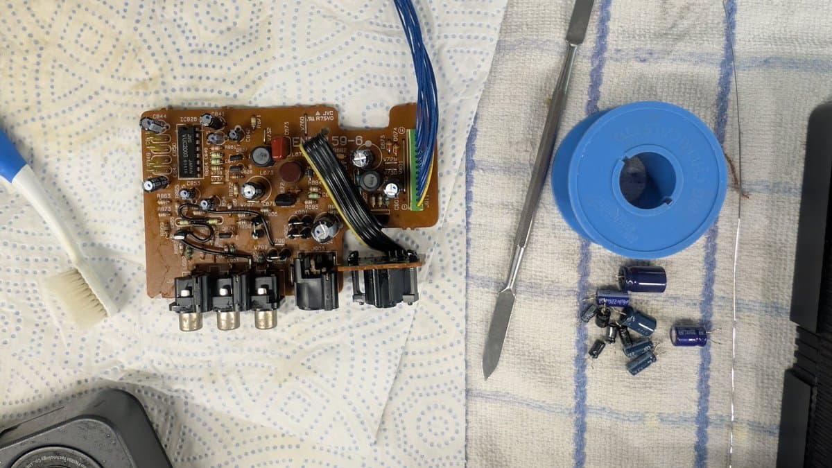 Disassembled Sega Wondermega video board with removed capacitors in blue spool, soldering iron, and IC chips laid out for recapping repair.
