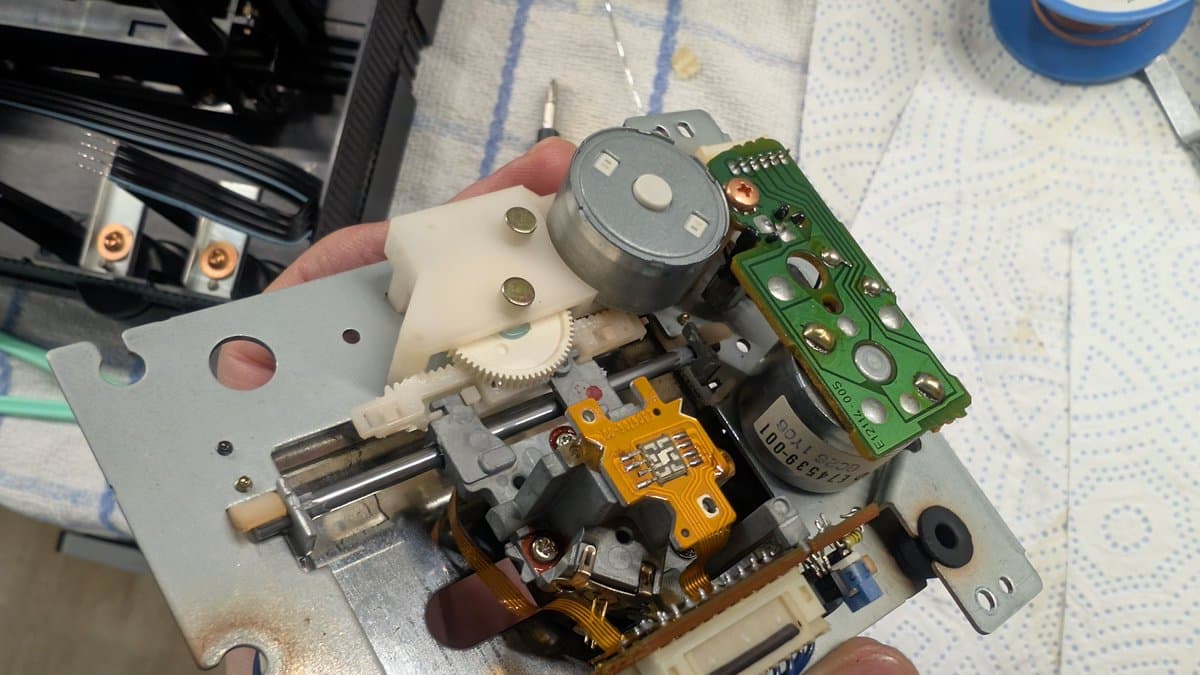 Wondermega laser PCB assembly held in hand showing recapped components, green control board, JVC Optima-5 pickup, gears, and sled mechanism during repair work.