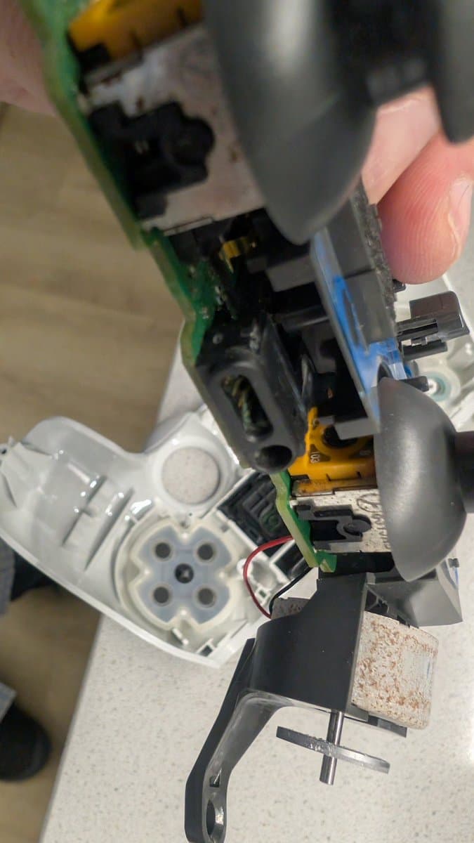 Close-up of PS4 controller internals during refurbishment; green PCB, black ribbon cable, corroded external port connector being cleaned with tool.