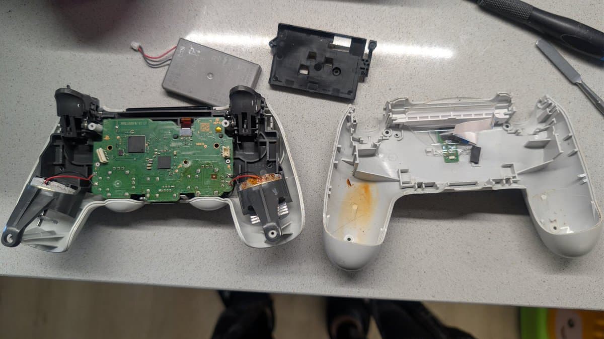 Disassembled PS4 controller internals showing green PCB, black buttons, and white housing halves; rusted rumble motors and battery cover visible above.