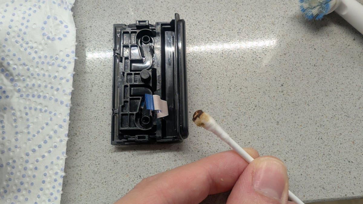 Disassembled PS4 controller showing internal components with a cleaning brush applicator positioned near the potentiometer; blue ribbon cable visible.
