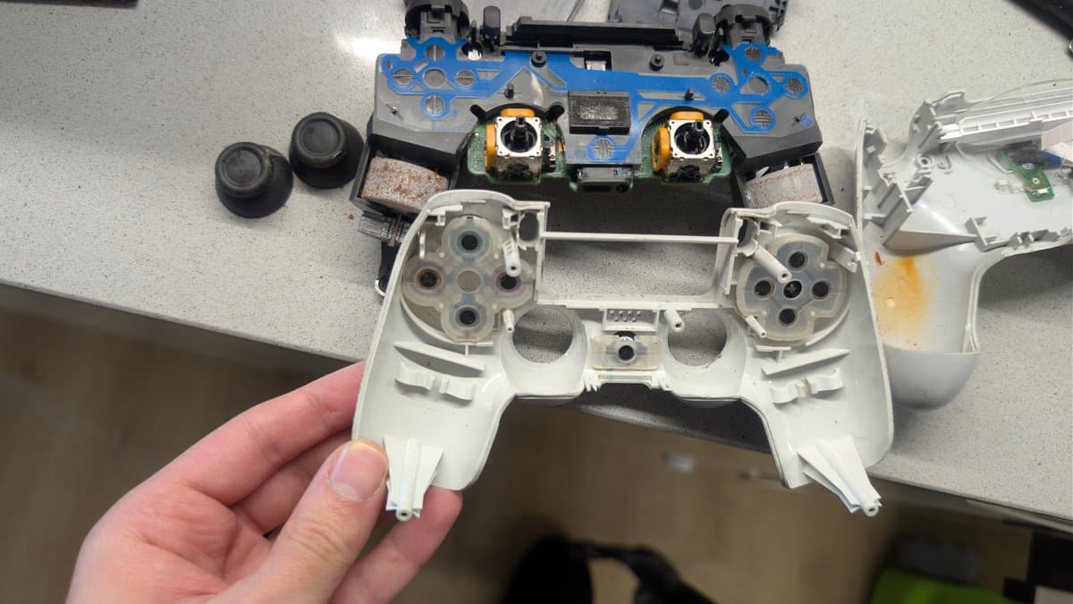 Disassembled PS4 controller internals: white shell held in hand, blue PCB with two joystick modules showing rust/corrosion, rumble motors, and separated components ready for ultrasonic cleaning.