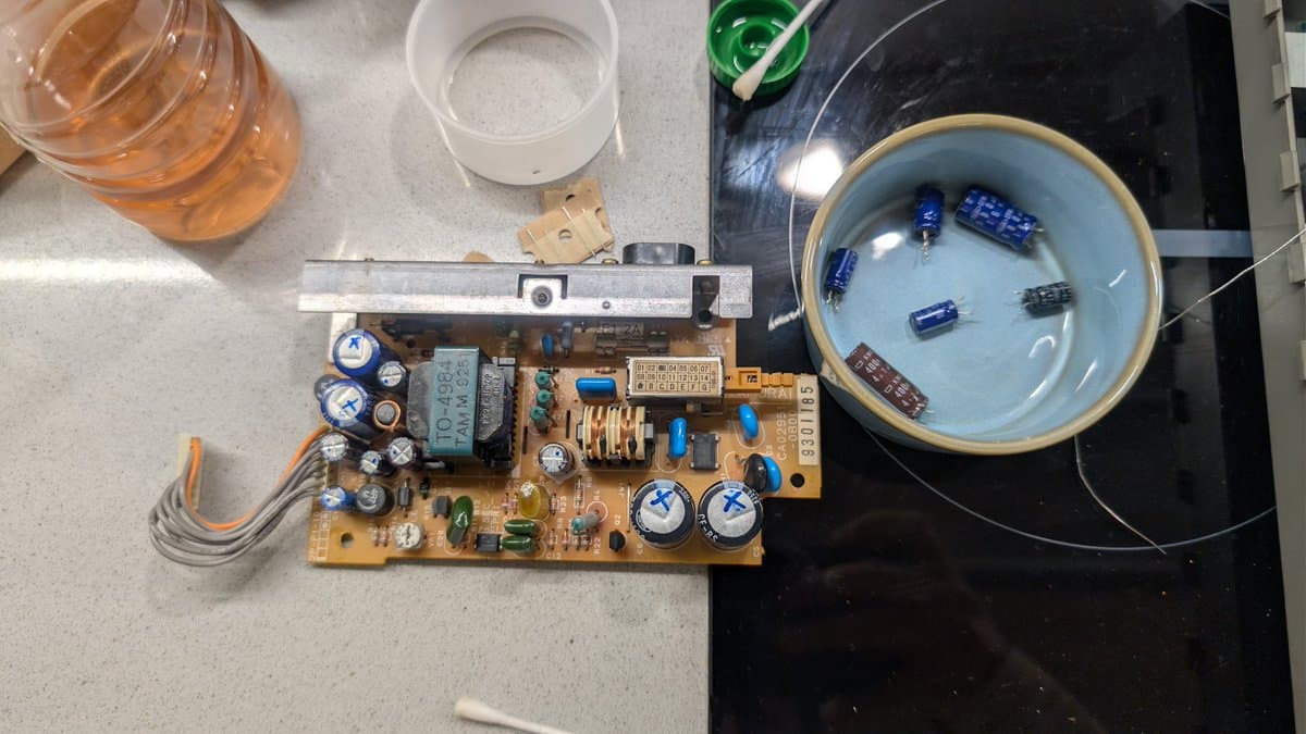 Fujitsu FM Towns Marty PSU board undergoing recap; electrolytic capacitors marked with X for replacement, bowl of desoldered components nearby.