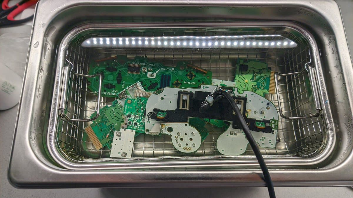 Multiple PCBs in ultrasonic cleaner basket with LED strip lighting, including green circuit boards and white component plates, mid-cleaning process.