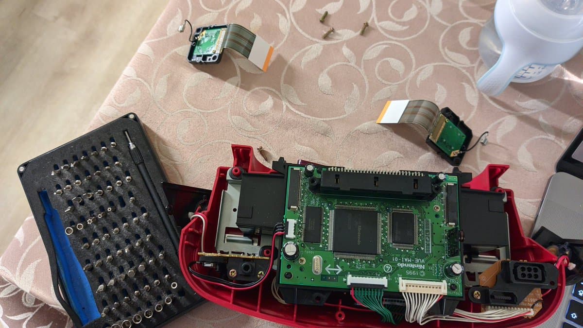 Disassembled Virtual Boy mainboard in red housing with flex cables, chip modules, bit driver set, and mounting jig during Virtual Ribbon 4.0 installation.