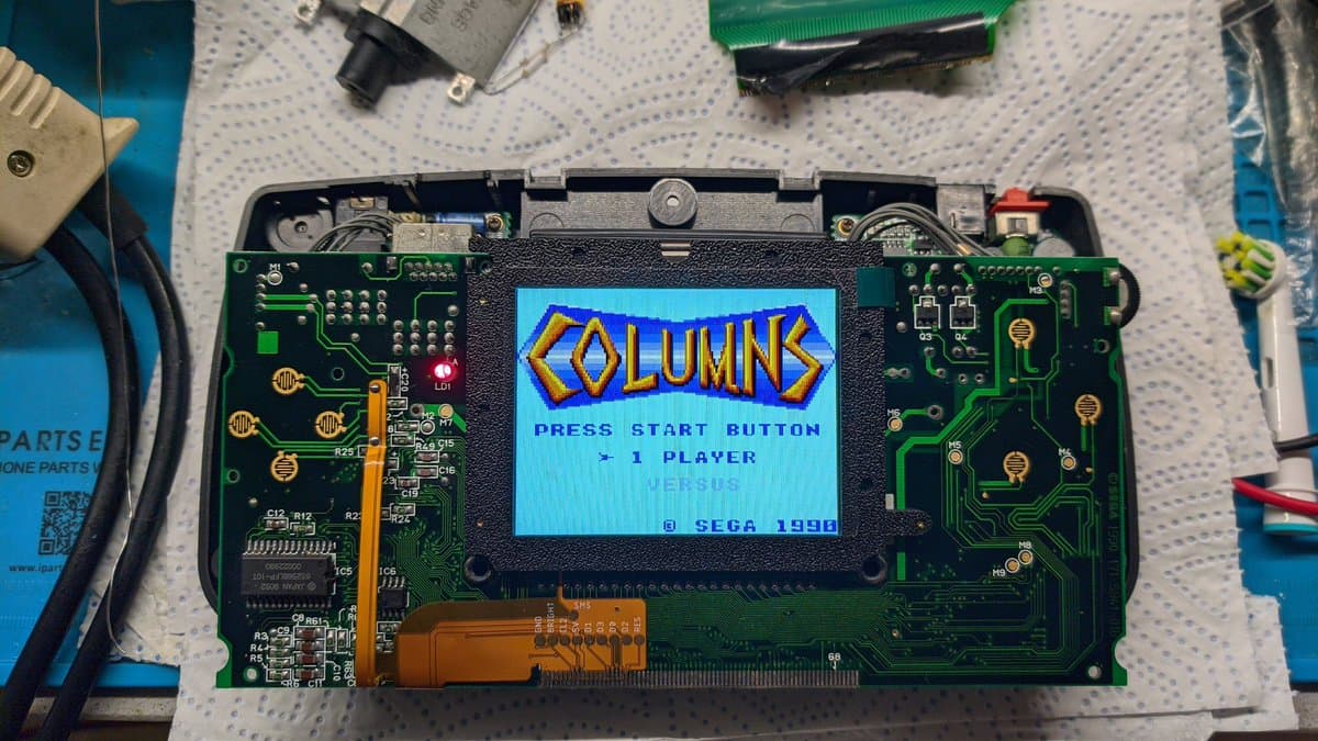 Sega Game Gear motherboard with IPS display installed showing Columns title screen, flex cable damage visible, clock line bodge wire repair evident on green PCB.