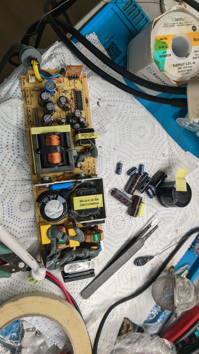 Disassembled Xbox 360 S PSU showing capacitors, transformers, and solder joints during recap and reflow repair work with tools nearby.