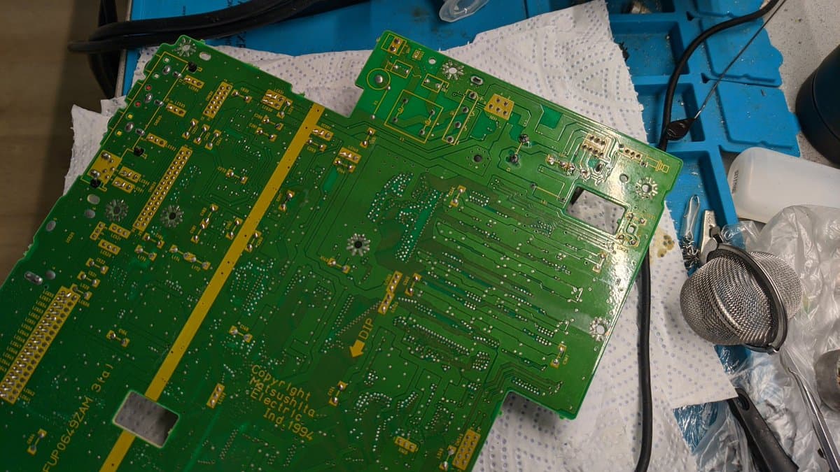 Panasonic FZ-1 motherboard mid-recap with corroded through-hole solder joints visible, soldering iron and solder sucker nearby on work mat.