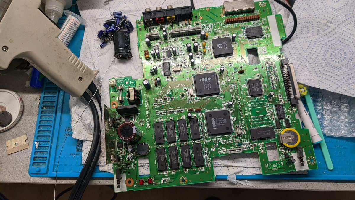 Panasonic FZ-1 mainboard during recap: green PCB with multiple ICs, reflowed solder joints, capacitor residue, cleaning tools, and blue work mat visible.
