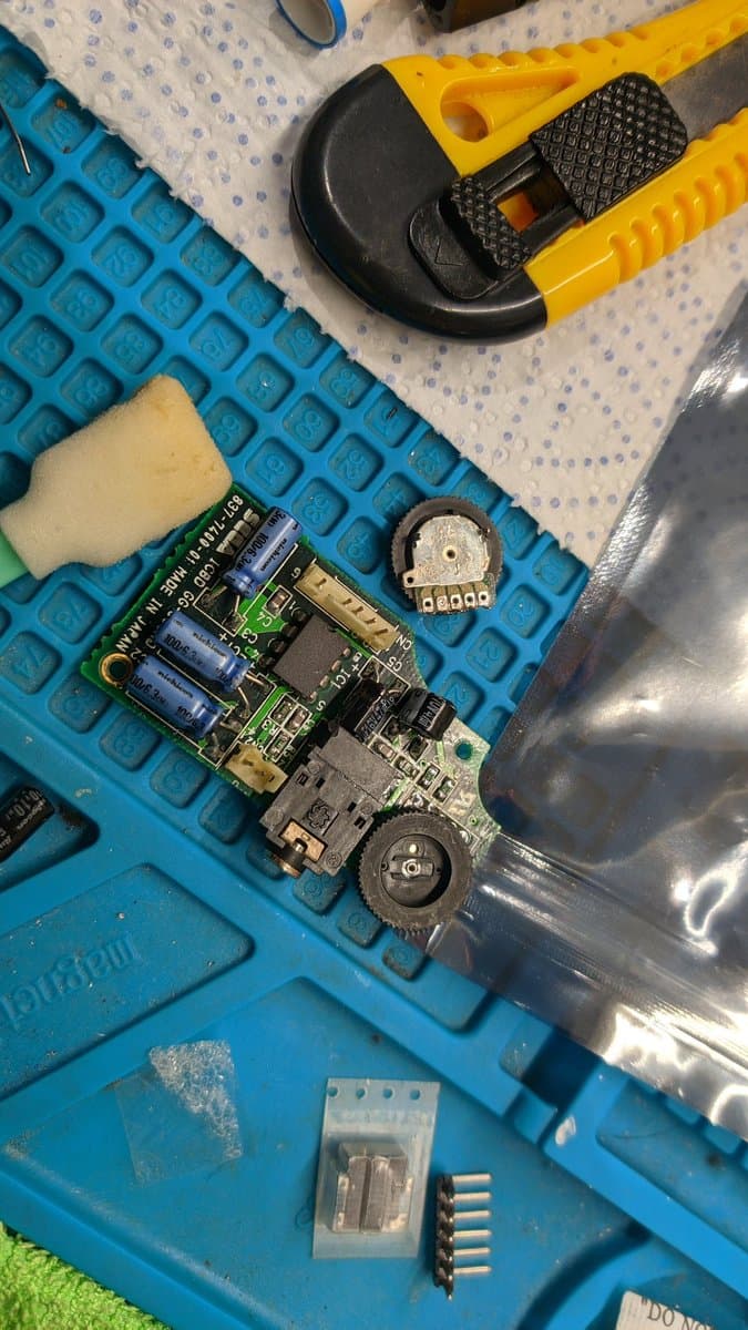 Game Gear circuit board with clock line bodge wire, capacitors, and potentiometer visible on blue work mat with tools and replacement parts nearby.