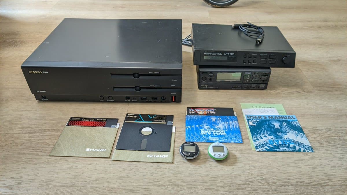 Sharp X68000 Pro II with Roland MT-32 and MT-102 synthesizers, floppy disks, manuals, and handheld games displayed on wooden surface.