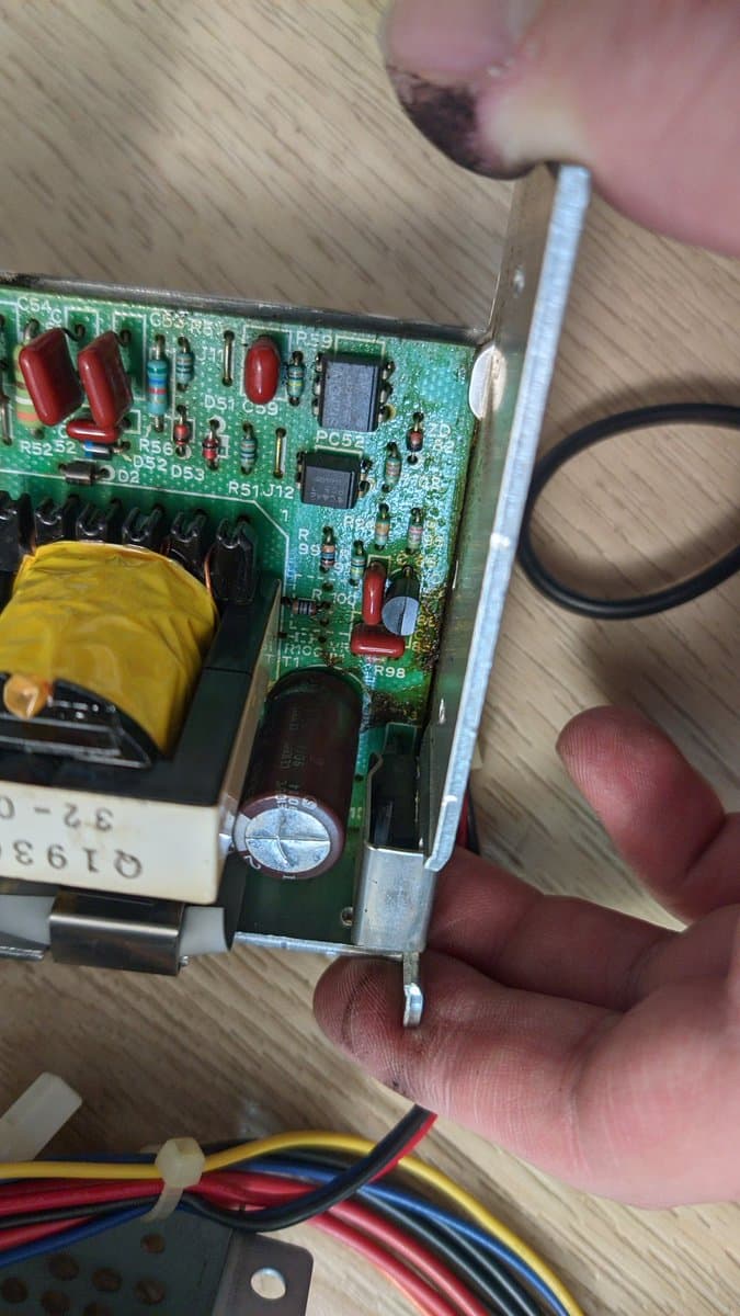 Hands holding Sharp X68000 Pro II power supply PCB showing corroded capacitor leakage residue, yellow transformer, and IC chips during restoration diagnostics.