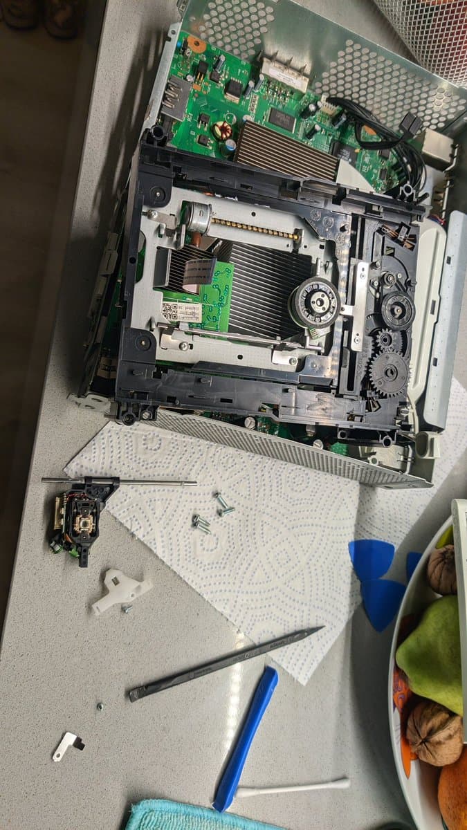 Xbox 360 disassembled showing DVD drive with removed laser assembly, heatsinks, circuit boards, and repair tools on work surface.