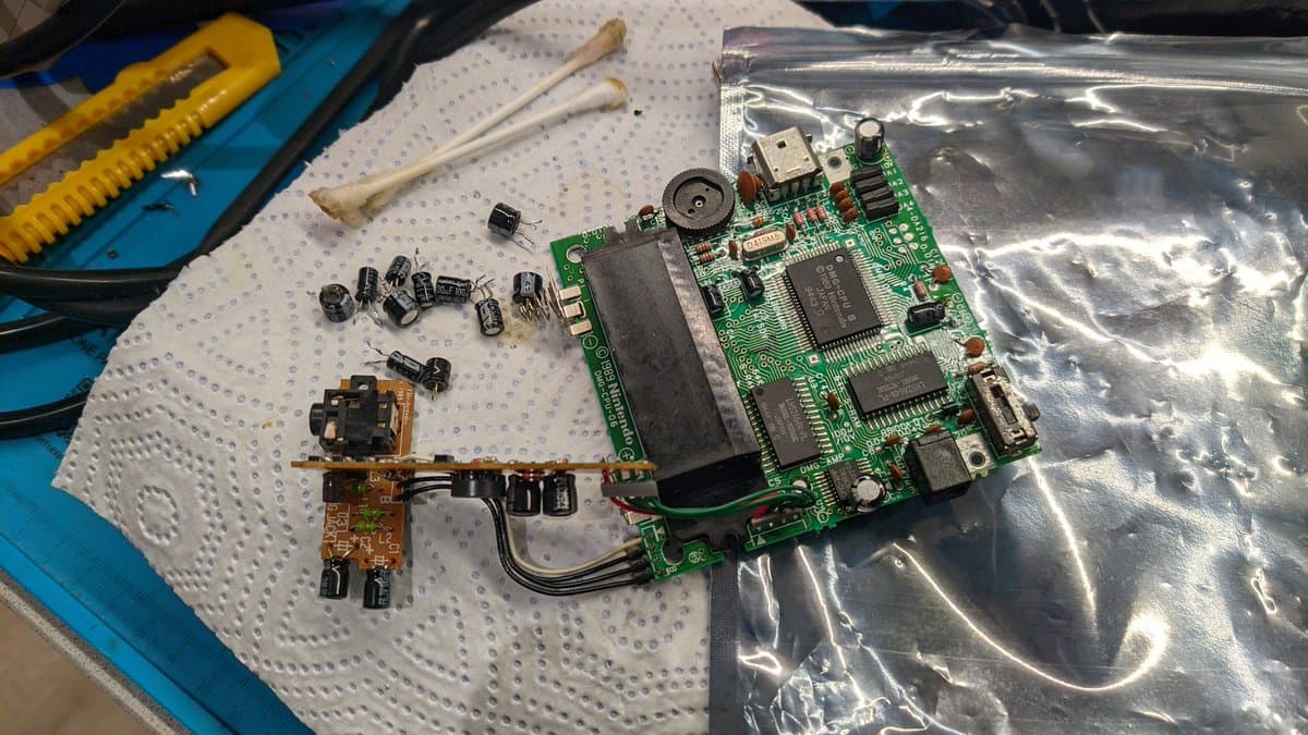 Disassembled Game Boy motherboard with removed capacitors, transistors, and power board during restoration work on white paper mat.