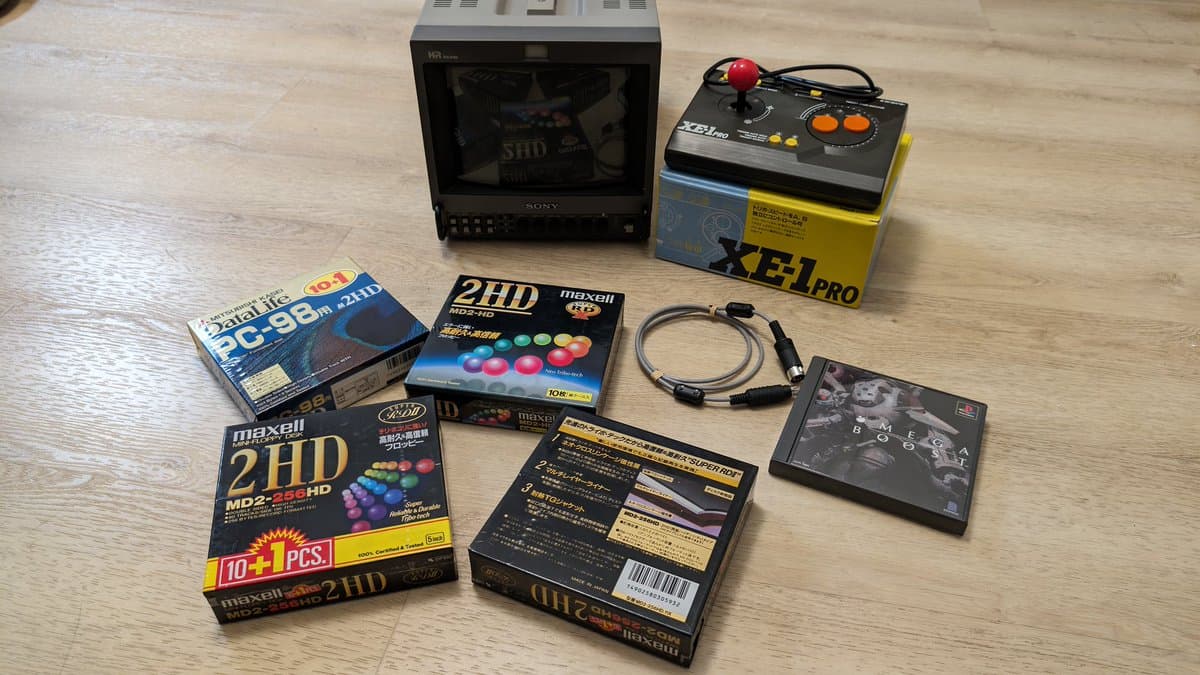 Sony PVM-9045QD CRT monitor displaying test pattern, with Maxell 2HD floppy disks, XEI Pro joystick, MIDI cable, and Omega Boost PS1 game on wooden surface.