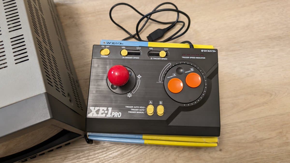 XEI Pro joystick controller with red ball top and orange buttons on wooden surface next to computer equipment.