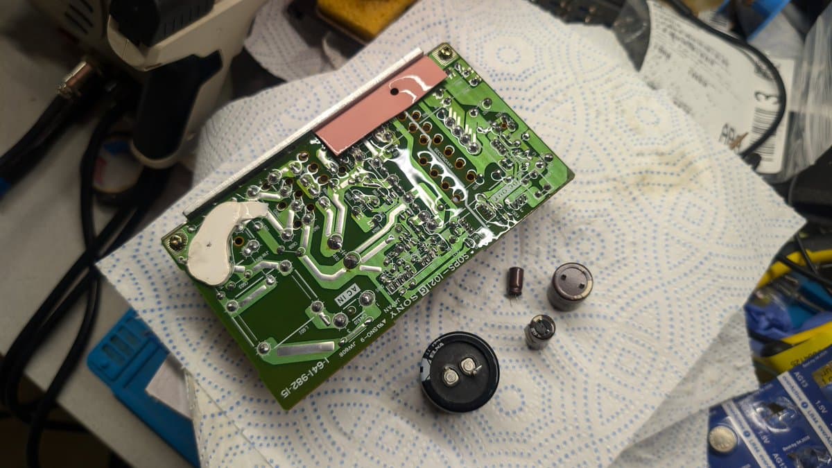 Green PCB with reflowed solder joints, removed capacitors and electrolytic component beside it on white work mat during PSU recap.