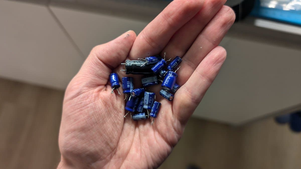 Hand holding a collection of blue electrolytic capacitors removed during X68000 partial recap, including what appears to be a black component, against an indoor background.