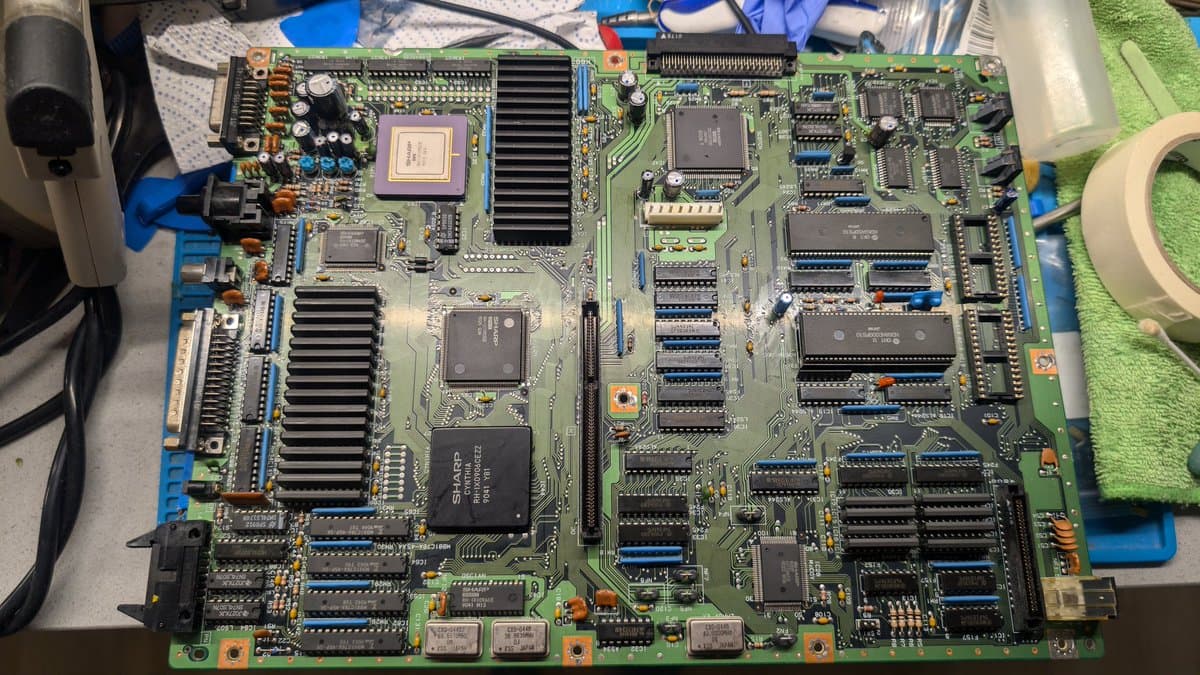 Sharp X68000 Pro II mainboard with multiple IC chips, heat-sinked components, and blue connector strips during partial capacitor replacement repair work.