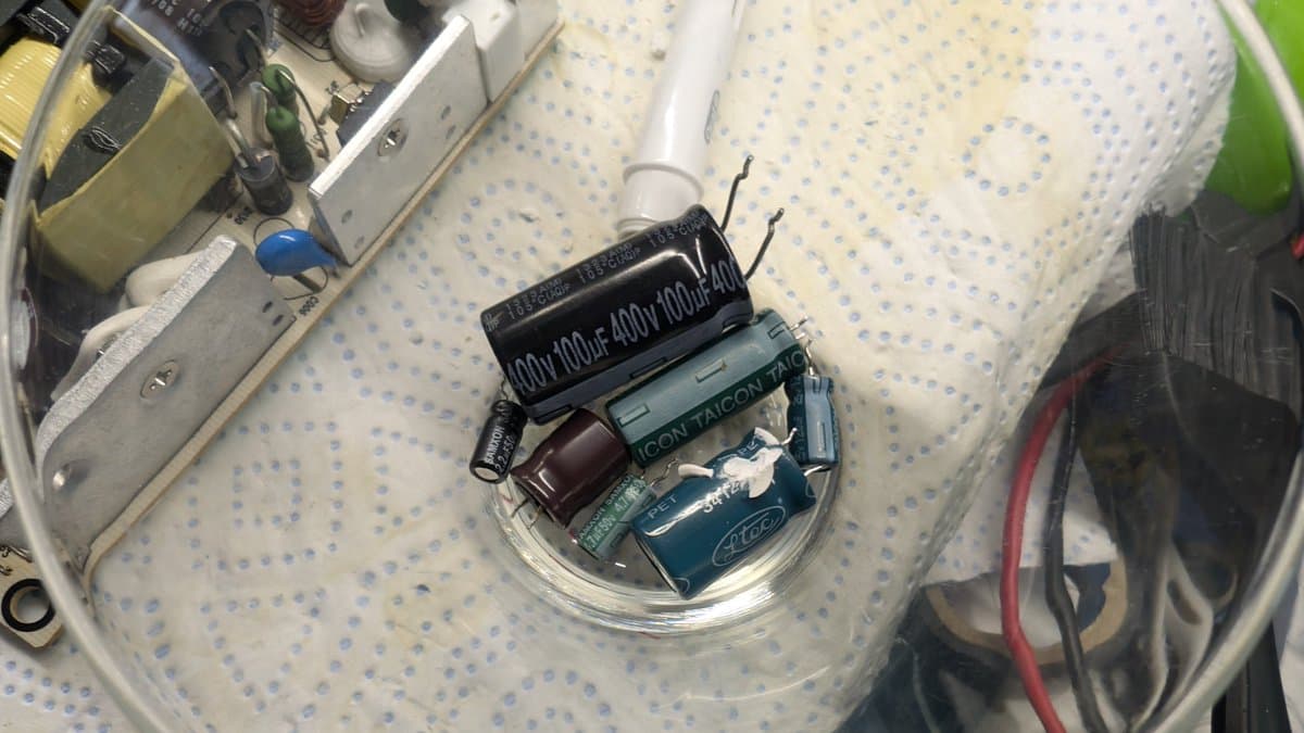 Close-up of Xbox 360 E PSU with old capacitors removed: black 100µ/400V Ltec cap, teal Taicon cap, brown capacitor in clear dish on dot-patterned surface.