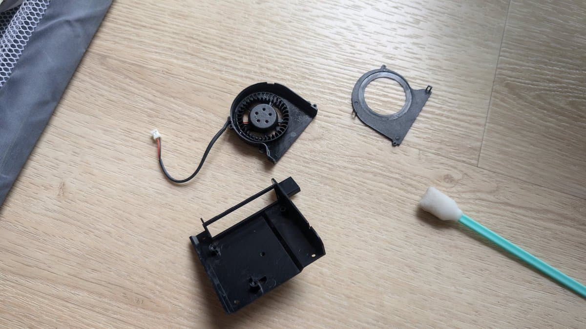 Disassembled Xbox 360 E PSU components: centrifugal fan with connector, shroud ring, housing, and cleaning brush on wooden surface.