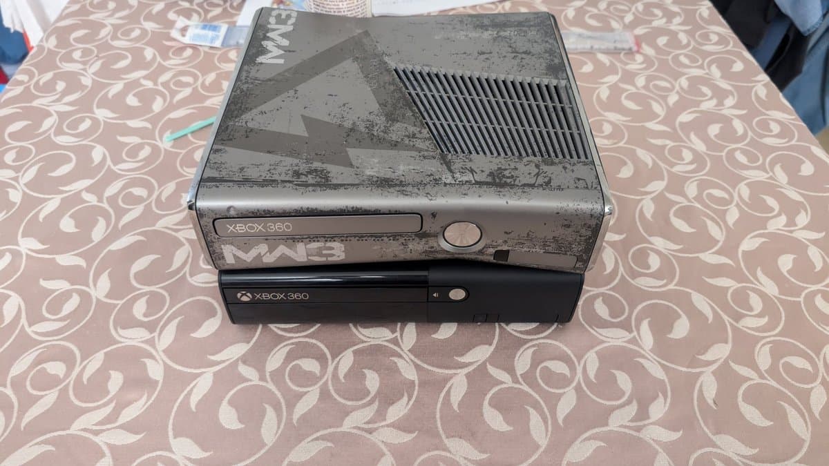 Two Xbox 360 consoles stacked on patterned fabric: dusty gray MW3 console on top, black console below. Both show Xbox 360 branding.