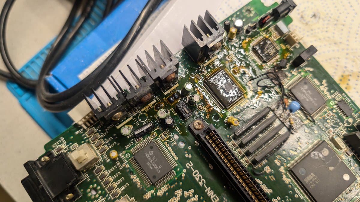 Corroded Sega Wondermega circuit board with leaking capacitor damage; multiple IC chips and heat sinks visible among oxidized solder joints.