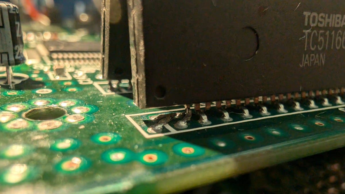 Close-up of Wondermega MCB showing damaged RAM package with broken legs next to IC chip, green PCB with solder joints and copper pads visible.