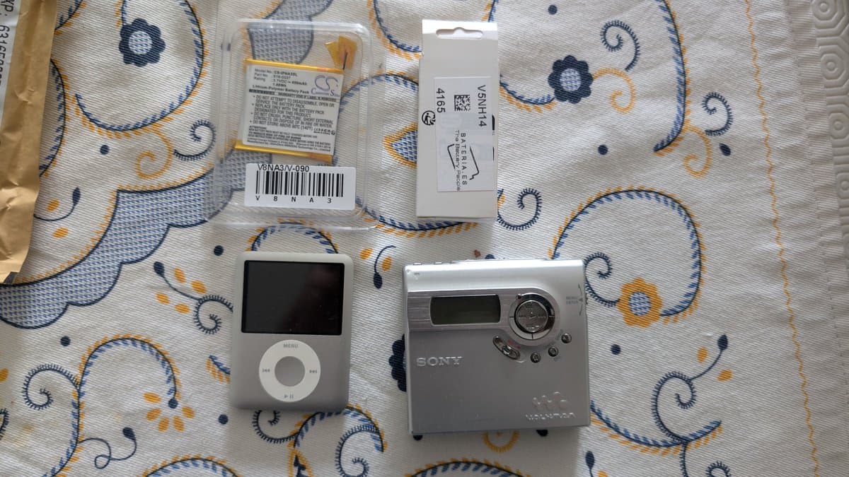 iPod Nano and Sony MZ-N920 MiniDisc Walkman with replacement batteries and packaging on decorative fabric.