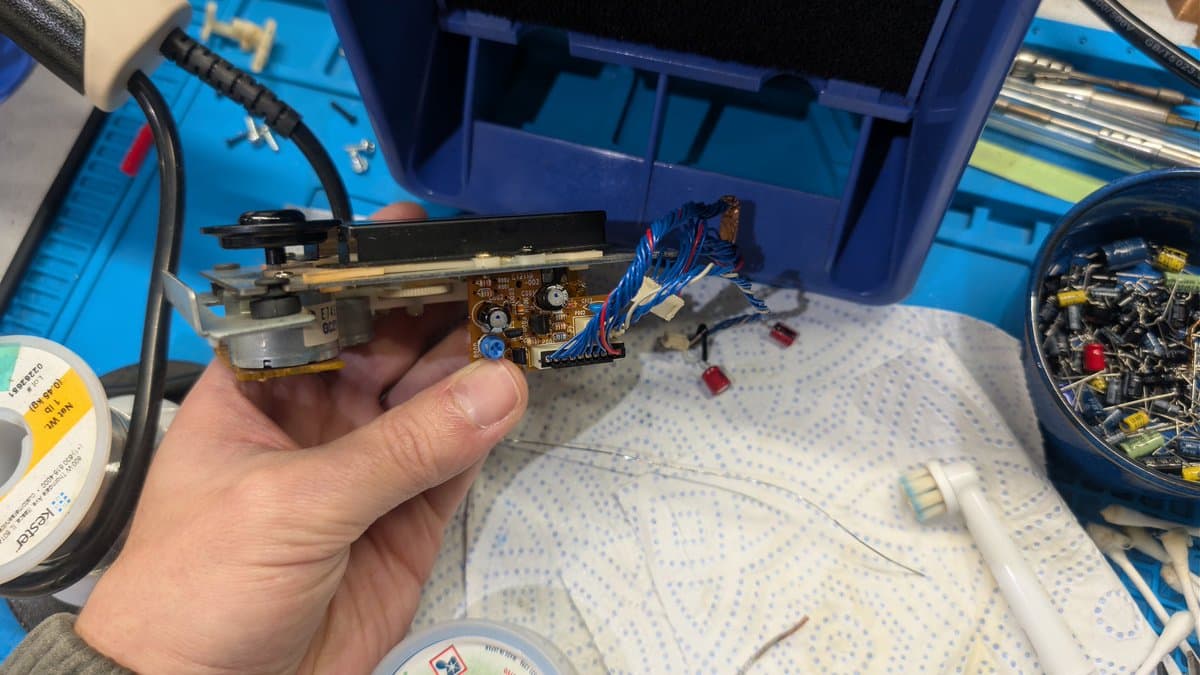 Technician holding Sega Wondermega audio/karaoke effects board with motor assembly and blue wiring harness over workbench during restoration.