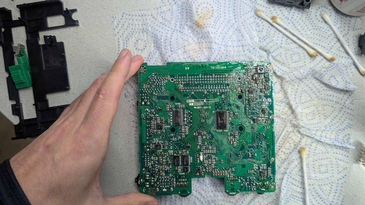 Hand holding NEC PC Engine LT mainboard after vinegar treatment, showing heavy capacitor leakage damage on component-dense green PCB.