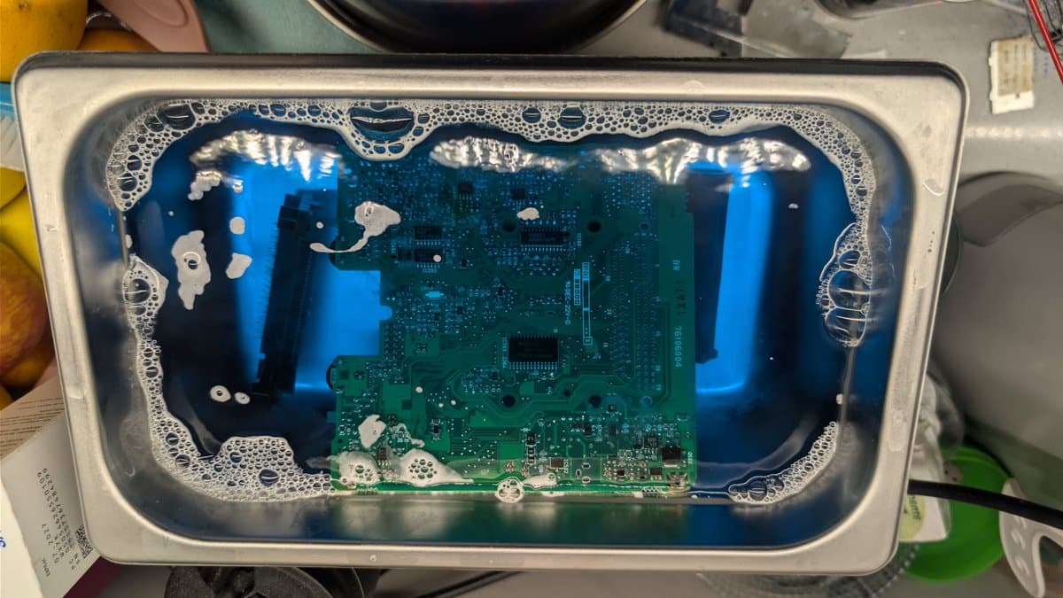 NEC PC Engine LT circuit board after ultrasonic cleaning, showing green PCB with blue capacitors and white mineral deposits around edges in clear plastic container.
