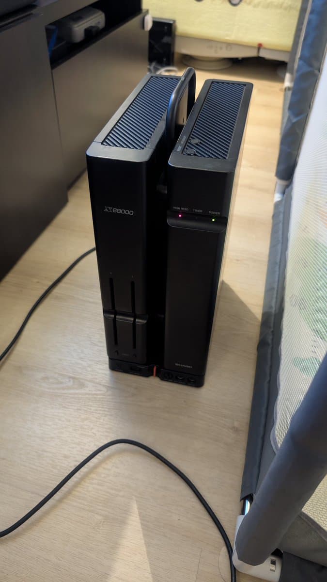 Two black tower cases on wooden surface; left unit labeled X68000, right unit showing power and token indicators; repair attempt with VGA adapter and OSSC.