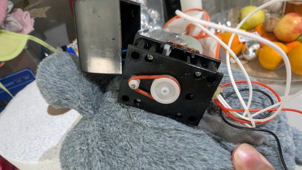 Close-up of Totoro toy mechanism showing black plastic gear assembly with white and orange gears, foam padding, and red/white wiring on gray fabric surface.