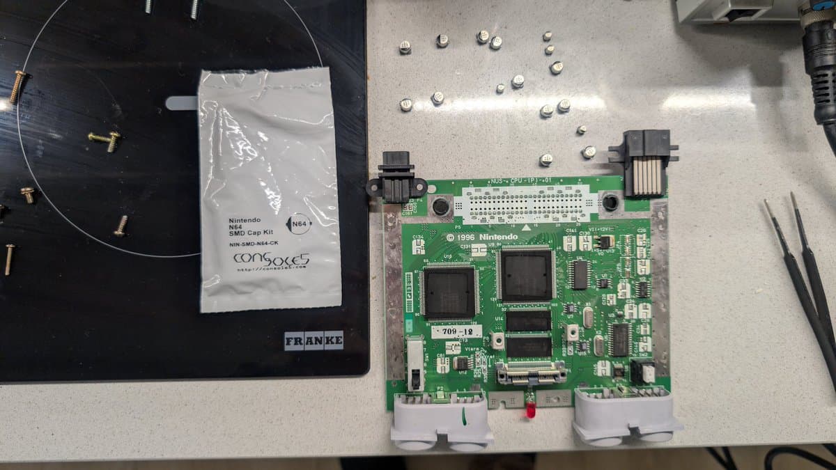 Nintendo 64 motherboard with capacitors removed during recapping repair, alongside SMD cap kit and organized replacement capacitors on work surface.