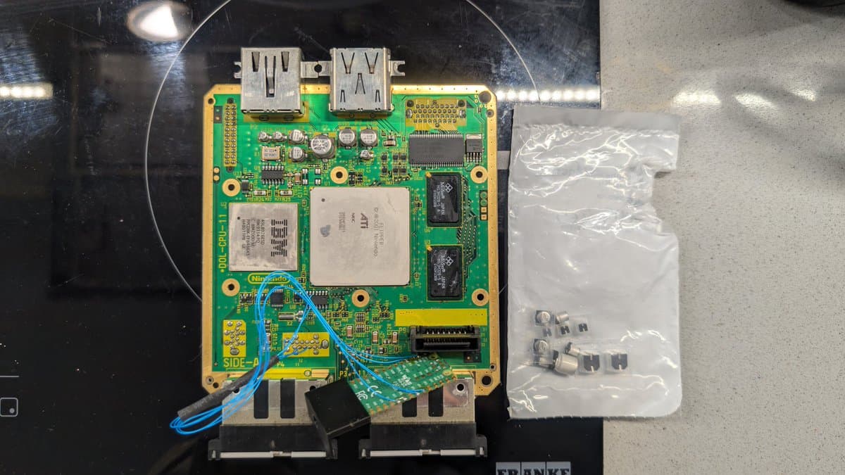 GameCube motherboard with USB ports, CPU heatsink, RAM chips, and capacitors visible; small components in plastic bag beside board.