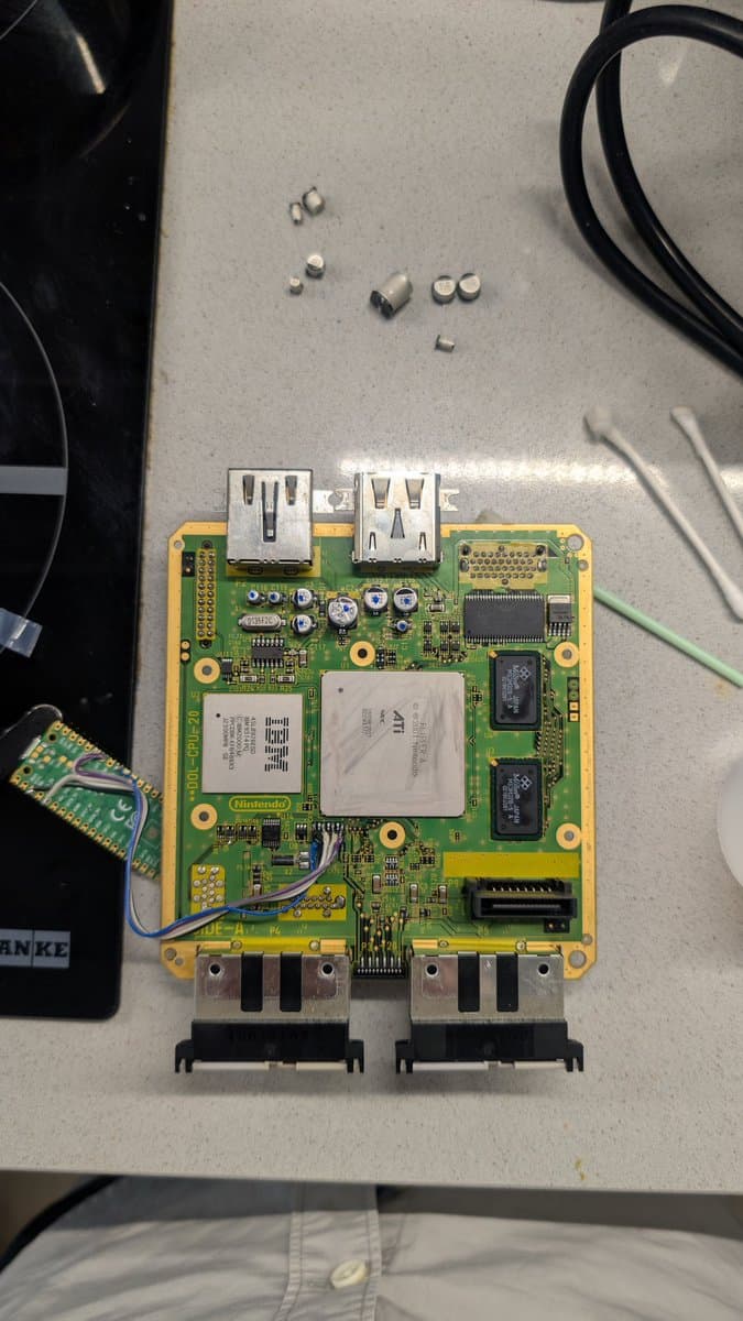 GameCube motherboard with two USB ports, recapped capacitors, and RAM chips visible after restoration work. Small components scattered above board.