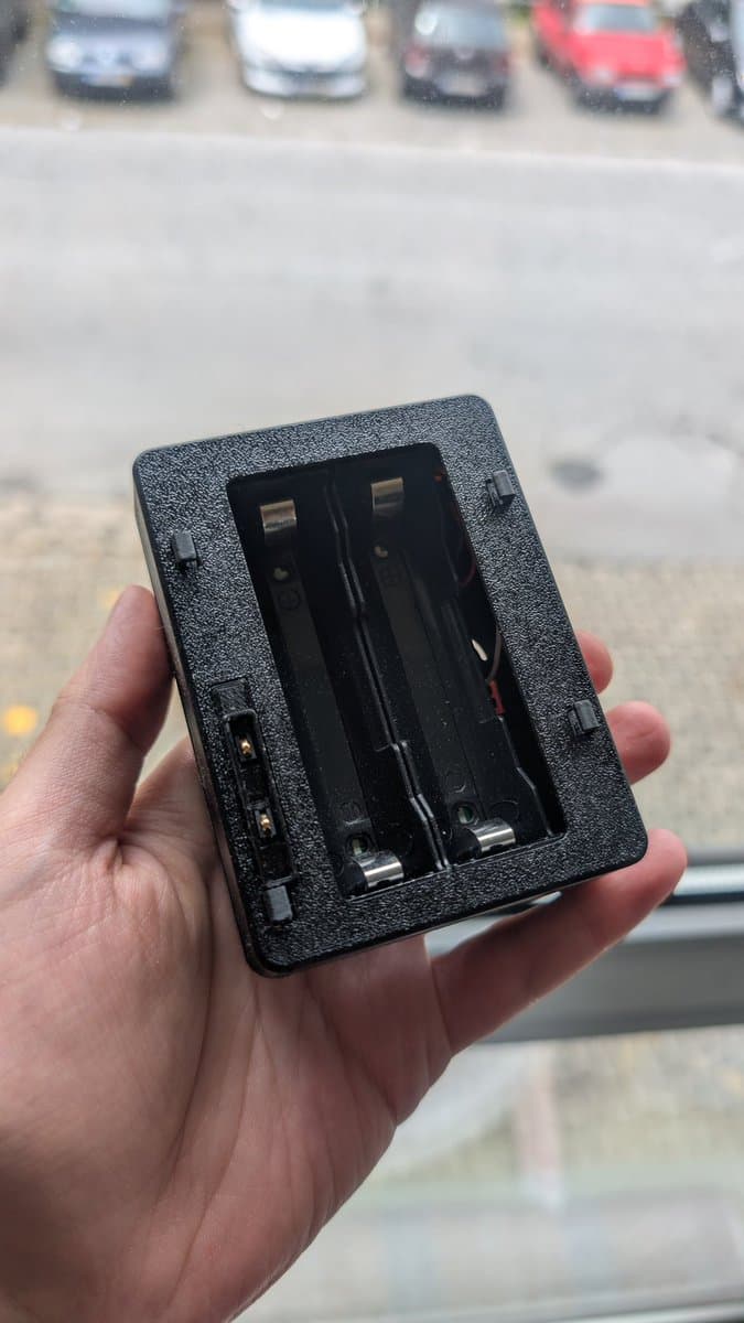 Hand holding black plastic Nomad battery pack with two battery compartments and metal contacts visible against parking lot background.