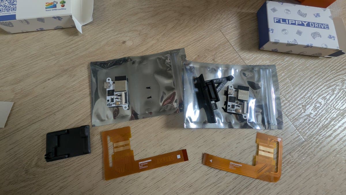 Panasonic Q optical drive components laid out: two anti-static bags containing circuit boards, two orange 3D-printed rail replacements, and one black plastic part on wooden workbench.