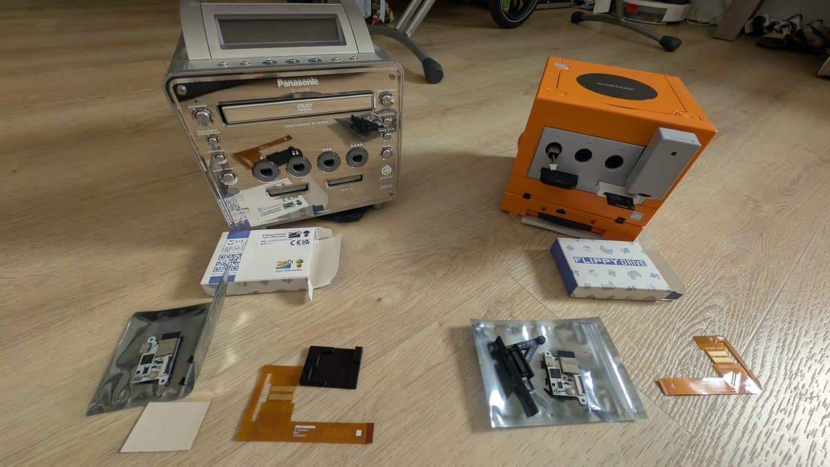 Disassembled spice orange GameCube next to Panasonic Q with FlippyDrive units, capacitors, and internal components laid out on workbench.