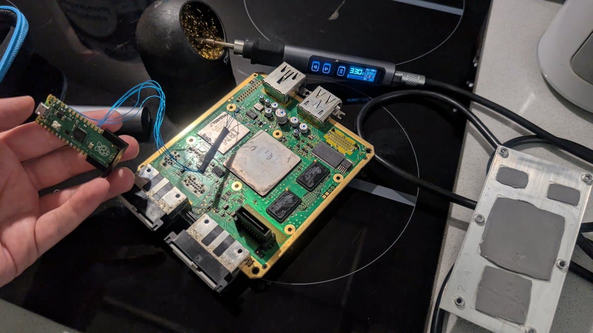 Technician holding GameCube motherboard with Picoboot installed alongside FlippyDrive board during swap installation, soldering iron visible.