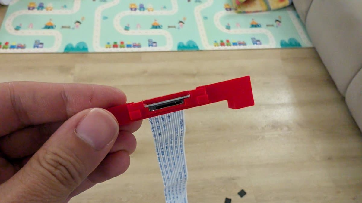 Red 3D-printed SD card extender with trimmed top piece held in hand, showing internal connector mechanism and blue ribbon cable attachment.