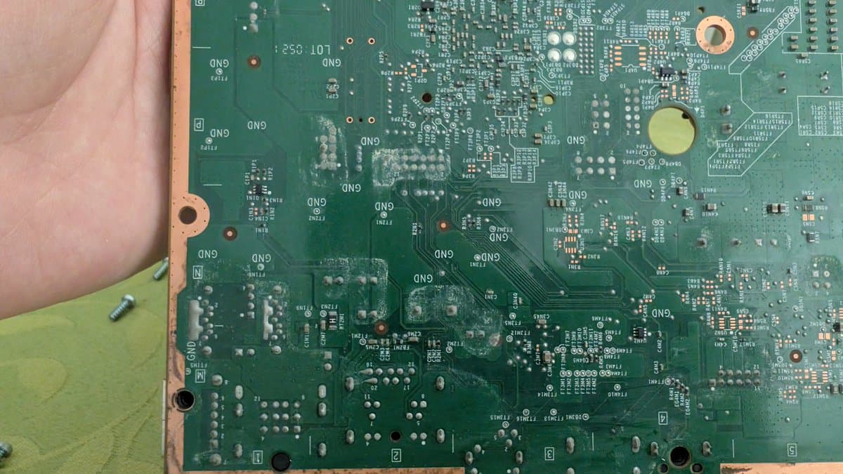 Xbox 360 E motherboard showing corrosion damage from liquid spill, with visible residue on circuit board and components requiring cleaning and repair.