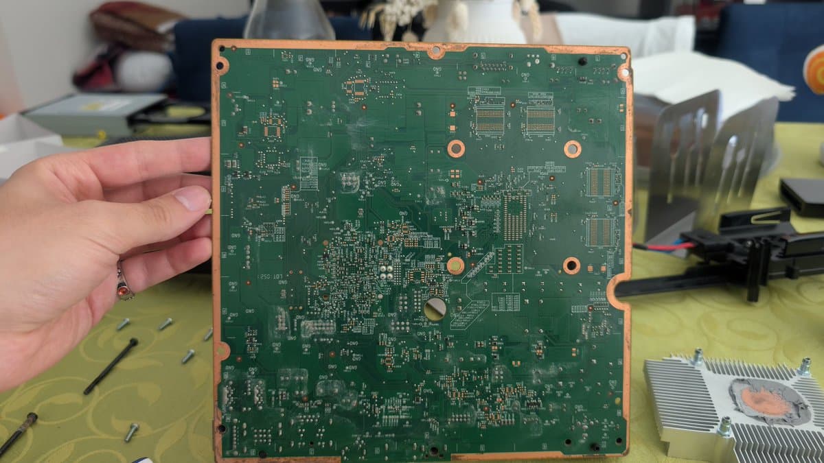 Xbox 360 E motherboard held in hand showing component-dense PCB with visible corrosion and discoloration from liquid damage; capacitors and solder joints visible.