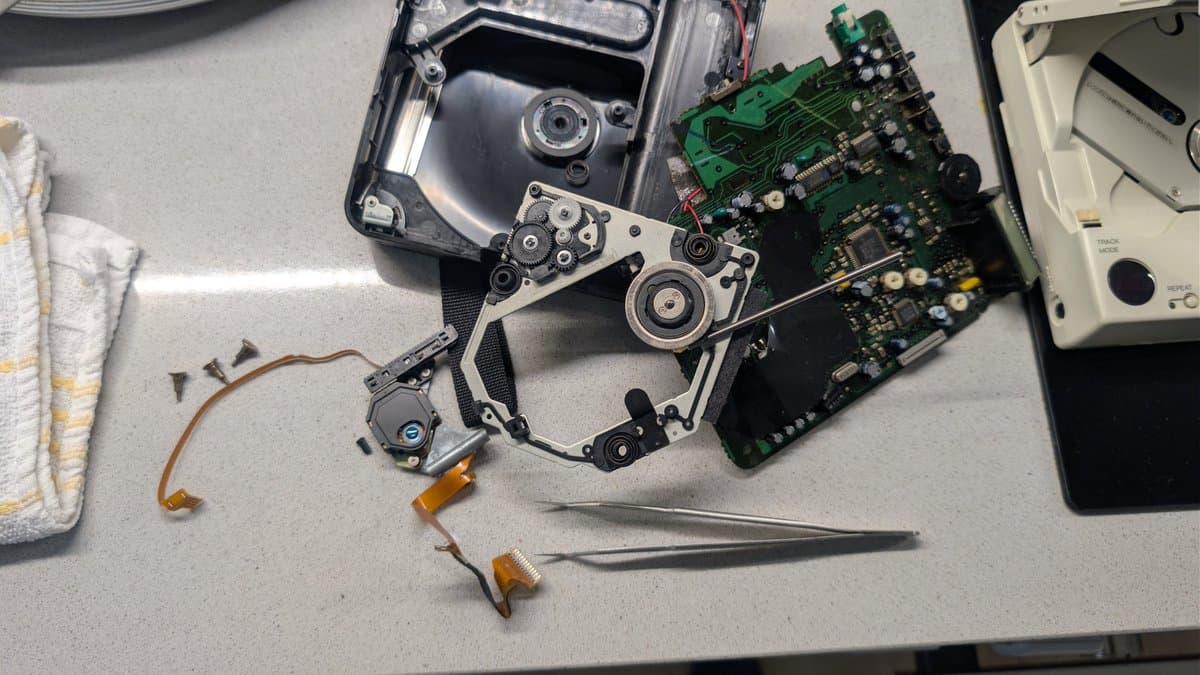 Disassembled PC Engine CD-ROM2 drive showing laser pickup mechanism, circuit boards, gears, and tweezers on work surface during restoration.